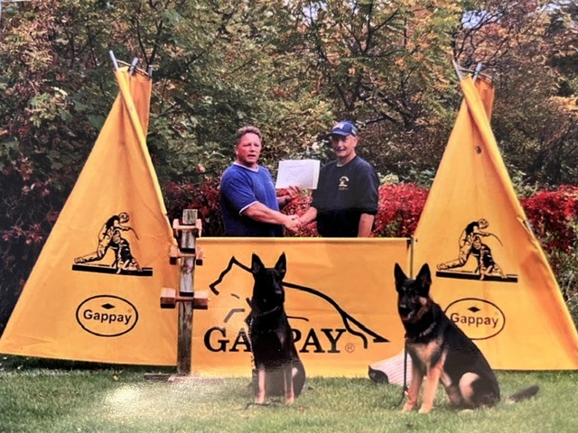 Two men shake hands in front of a Gappay banner, celebrating a dog training achievement. Two German Shepherds sit attentively nearby.