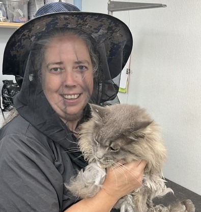 A groomer wearing a protective hat holds a fluffy gray cat in a grooming salon, showcasing pet care services.